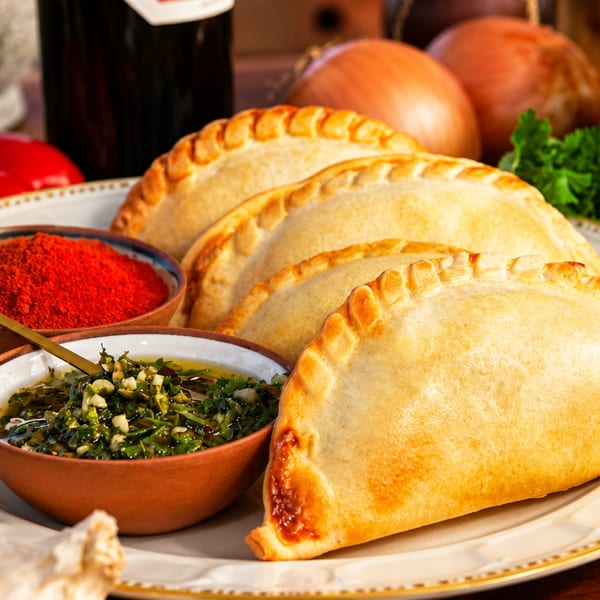 Golden-brown empanadas on a white plate with chimichurri, paprika, and fresh vegetables.