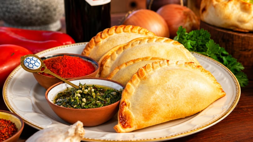 Golden-brown empanadas on a white plate with chimichurri, paprika, and fresh vegetables.