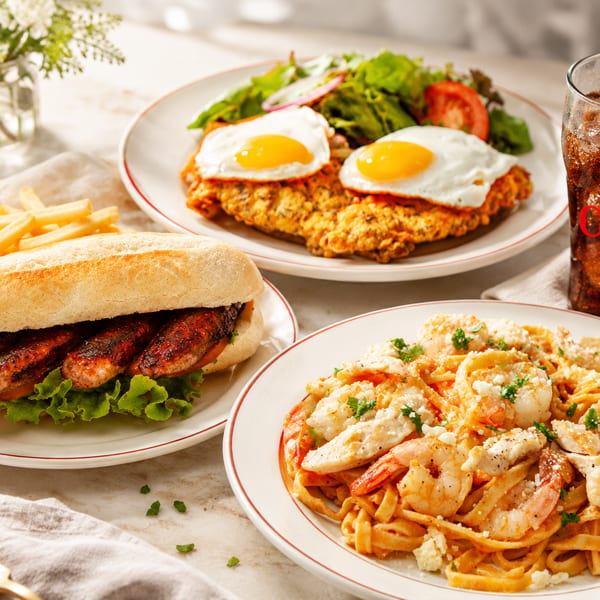 Three plates of food: a sausage sandwich with fries, chicken schnitzel with eggs and salad, and shrimp pasta.