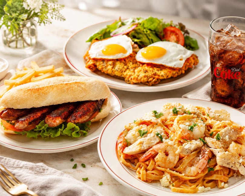 Three plates of food: a sausage sandwich with fries, chicken schnitzel with eggs and salad, and shrimp pasta.