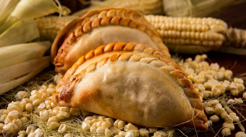 Three golden-brown empanadas with crimped edges rest on corn kernels and husks.