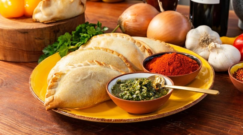Empanadas on a yellow plate with chimichurri, paprika, onions, garlic, and a bottle of wine.