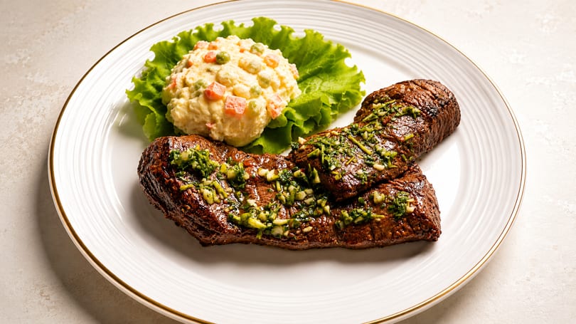 Grilled steak with chimichurri, potato salad, and lettuce on a white plate.