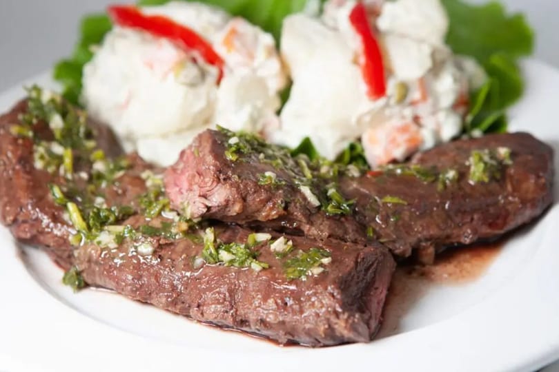 Grilled steak with chimichurri sauce, served with potato salad and red pepper garnish.