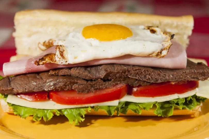 Hearty sandwich with steak, ham, fried egg, tomato, cheese, and lettuce on a baguette.