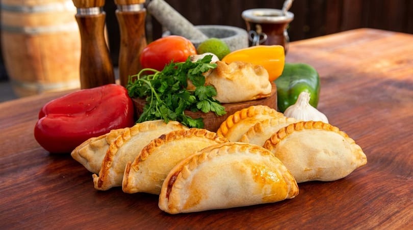 Golden-brown empanadas arranged on a wooden table with fresh vegetables and spices in the background.