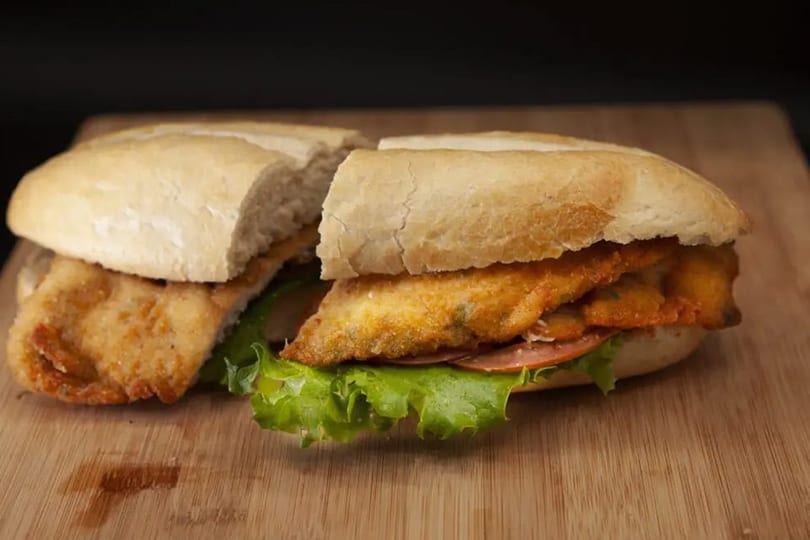 Crispy fried chicken sandwich with lettuce and tomato on a toasted bun, cut in half on a wooden board.