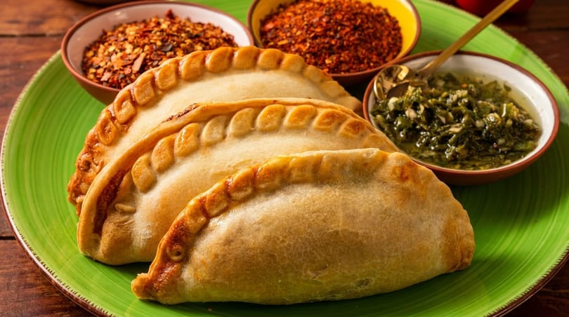 Three golden-brown empanadas on a green plate, with bowls of chili flakes and chimichurri sauce.