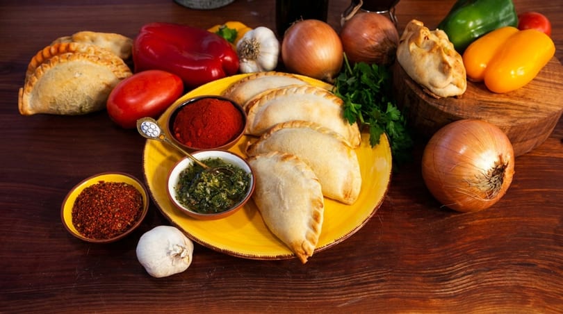Empanadas on a yellow plate with spices, fresh vegetables, and herbs on a wooden table.