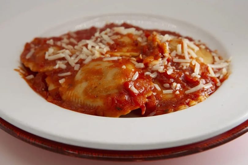 Close-up of ravioli covered in red sauce and sprinkled with white shredded cheese on a white plate.