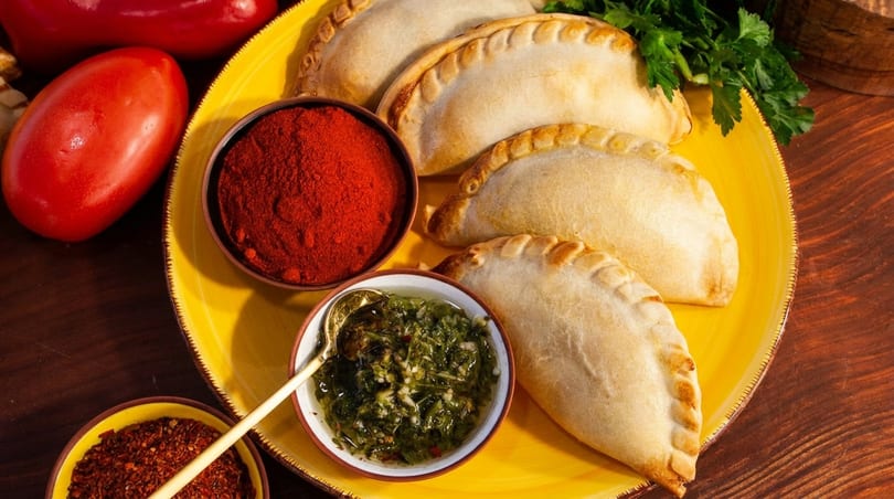 Empanadas on a yellow plate with red spices, green chimichurri sauce, and fresh vegetables.