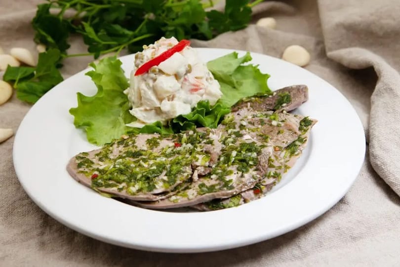 Sliced meat with green herb sauce, a side of potato salad, and lettuce on a white plate.