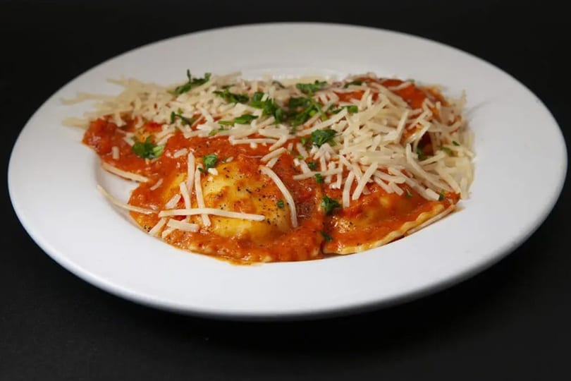 Plate of ravioli covered in red sauce, topped with shredded cheese and green herbs.